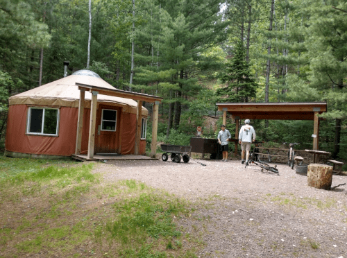 A yurt in a forested area with two people, a picnic table, and a bike nearby. Green trees surround the scene.