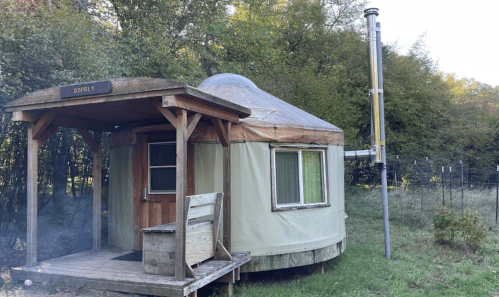 A cozy yurt with a wooden porch, surrounded by trees and a tall structure nearby, set in a natural landscape.