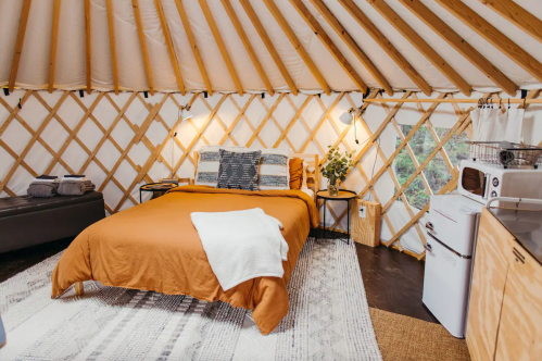 Cozy yurt interior featuring a warm orange bed, wooden accents, and a small kitchenette. Natural light filters in.