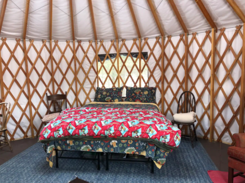Cozy yurt interior featuring a bed with a colorful quilt, wooden chairs, and a patterned rug.