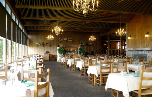 A spacious restaurant interior with wooden decor, tables set with white tablecloths, and chandeliers hanging from the ceiling.