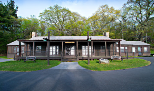 A rustic wooden building with a porch, surrounded by trees and a paved path, set in a natural landscape.