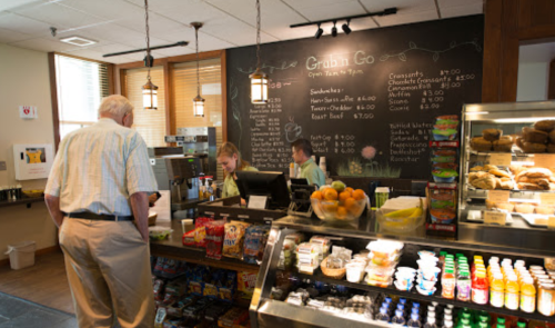 A customer at a café counter with staff, a menu board, and a display of snacks and drinks.