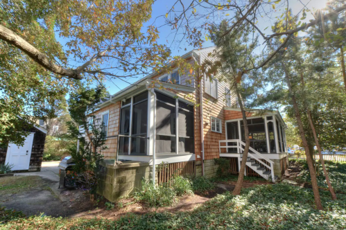 A charming two-story house with a screened porch, surrounded by trees and greenery on a sunny day.