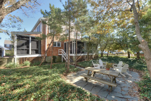 A cozy backyard with a stone patio, picnic table, and trees surrounding a charming house.