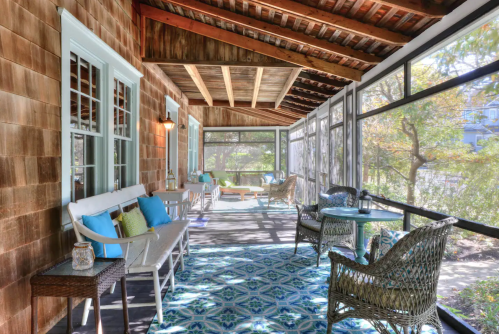 A cozy screened porch with wooden walls, comfortable seating, and a colorful rug, surrounded by greenery.