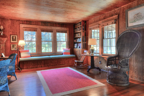 Cozy living room with wooden walls, large windows, a reading nook, and a wicker chair on a red rug.