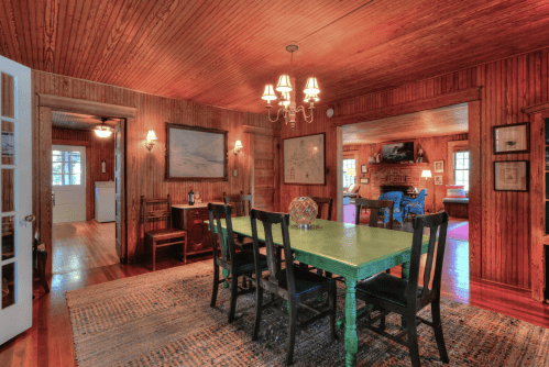 Cozy dining room with a green table, wooden walls, and warm lighting, leading to a living area.