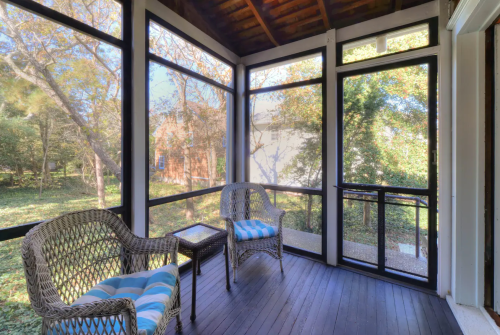 A screened porch with two wicker chairs and a small table, surrounded by trees and greenery.