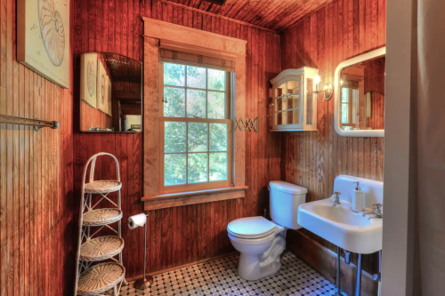 Cozy bathroom with wooden walls, a window, a sink, toilet, and a decorative shelf. Natural light fills the space.