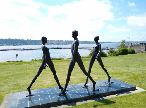 Three abstract bronze sculptures of figures walking, set against a scenic waterfront backdrop with blue skies.