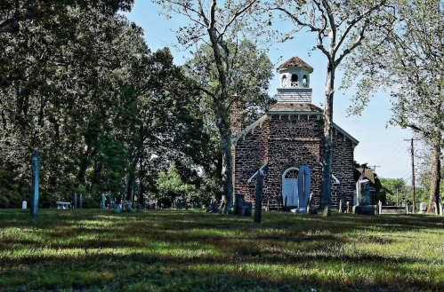A historic stone church surrounded by trees and gravestones, set in a peaceful, grassy cemetery.