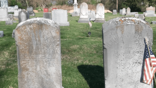 Two weathered gravestones in a cemetery, with small American flags placed at their bases, surrounded by green grass.