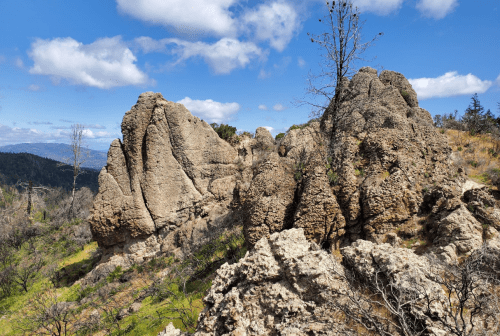 Rocky formations under a blue sky with scattered clouds, surrounded by sparse vegetation and distant hills.