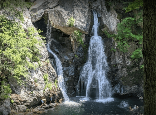 A serene waterfall cascades into a pool, surrounded by lush greenery and rocky cliffs. Two people explore the area.