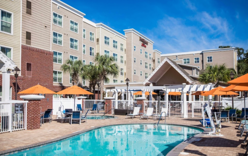 A hotel with a pool area featuring lounge chairs and orange umbrellas, surrounded by palm trees and blue skies.