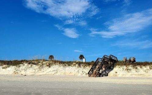 A wooden staircase leads from the beach to a sandy dune under a clear blue sky.
