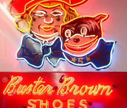 Neon sign featuring a girl and a dog, advertising "Buster Brown Shoes." Bright colors and playful design.