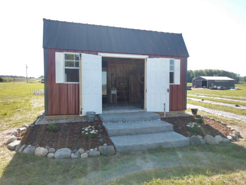 A red and white shed with open double doors, surrounded by a landscaped path and grassy fields.