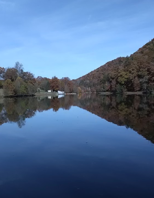 A serene lake reflecting trees and a blue sky, surrounded by autumn foliage and a distant white building.
