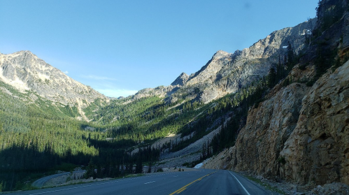 A scenic mountain road surrounded by lush green forests and rocky cliffs under a clear blue sky.