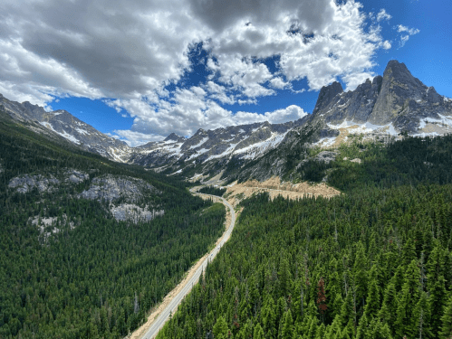 A winding road through lush green forests, surrounded by majestic mountains and a blue sky with fluffy clouds.