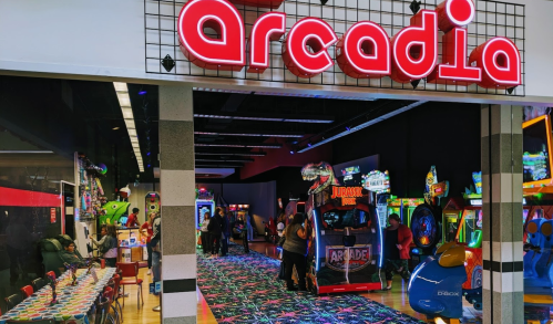 Brightly lit arcade entrance with "Arcadia" sign, featuring various games and people enjoying the space.