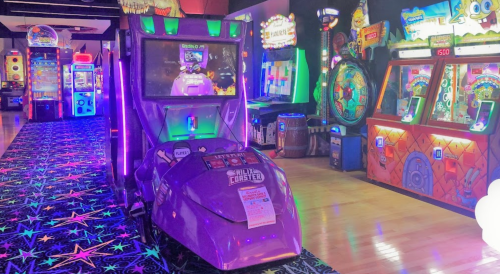 A vibrant arcade scene featuring a purple racing game machine surrounded by colorful lights and various arcade games.
