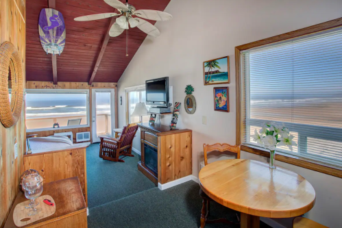 Cozy beachside living area with a wooden table, rocking chair, and ocean view through large windows.