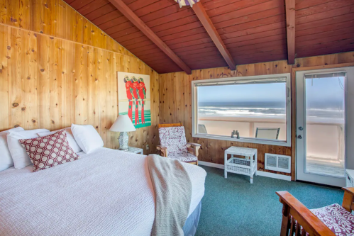 Cozy beachside bedroom with wooden walls, large window, and ocean view. Features a king bed and vintage armchairs.