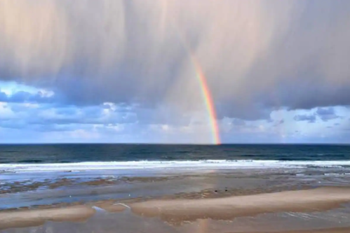 A vibrant rainbow arcs over a calm ocean, with sandy beach and dramatic clouds in the background.