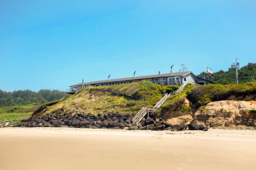 A beachside building on a grassy cliff, with stairs leading down to the sandy shore and rocky coastline.