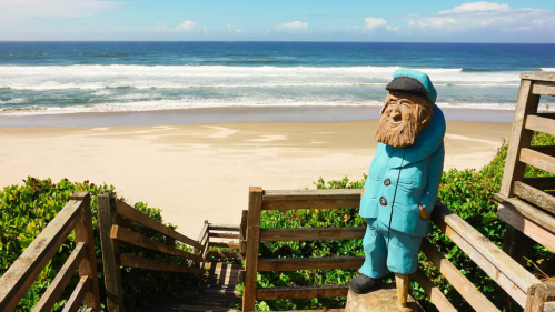 A wooden statue of a man in a blue coat stands on a staircase overlooking a sandy beach and ocean waves.