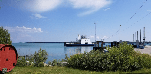 A ferry approaches a dock on a calm lake, surrounded by greenery and under a clear blue sky.