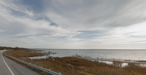 A scenic view of a calm lake with grassy shores under a cloudy sky, alongside a winding road.
