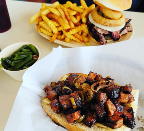 A sandwich with diced meat and onions on a bun, served with green beans and a plate of crinkle-cut fries in the background.