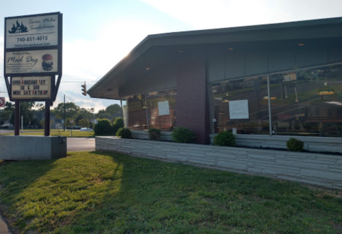 A sandwich shop with a sign advertising breakfast and lunch, featuring large windows and a grassy area in front.