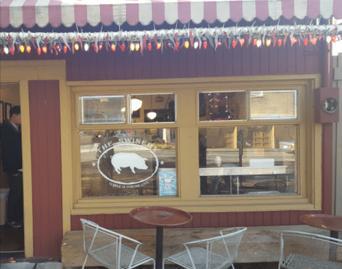Exterior of a restaurant called "The Swine," featuring a festive awning and outdoor seating.