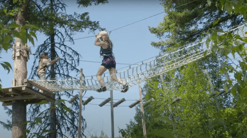 A person crosses a rope bridge in a forested area, while another individual observes from a platform nearby.