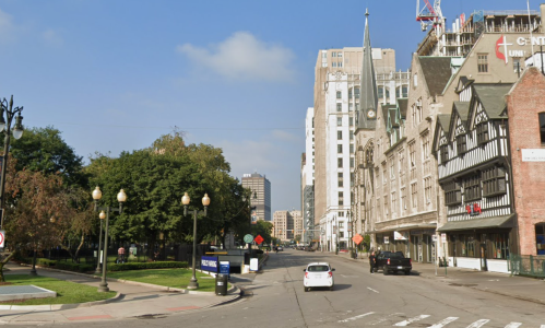 A city street with trees, buildings, and a clear blue sky, featuring a mix of modern and historic architecture.