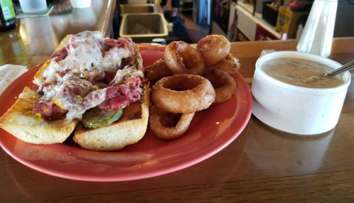A plate with a sandwich topped with meat and cheese, onion rings, and a bowl of soup on a wooden table.