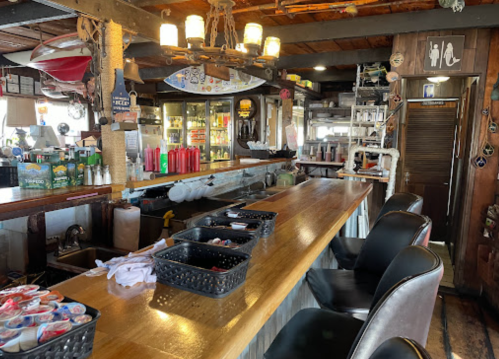A rustic bar interior with wooden counters, black chairs, and shelves stocked with drinks and condiments.