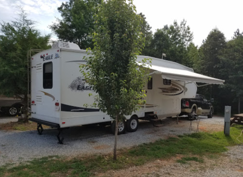 A parked RV with an awning, surrounded by trees and gravel, in a campsite setting.