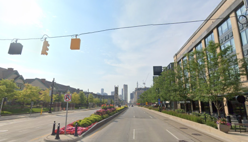 A city street lined with trees and flowers, featuring traffic lights and buildings in the background under a clear sky.