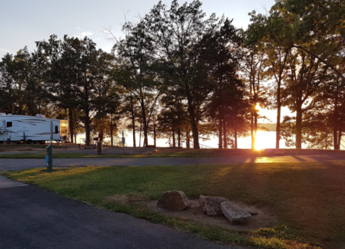 A serene campsite at sunset, with an RV parked among trees and a view of the sun setting over the water.