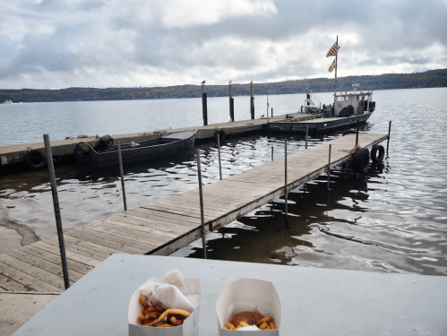 A wooden dock extends into a calm lake, with a boat moored nearby and takeout containers in the foreground.