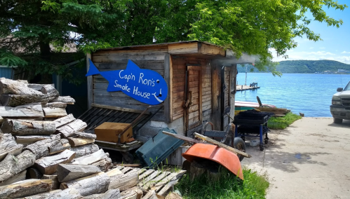 A rustic smokehouse with a blue sign reading "Cap'n Ron's Smoke House," surrounded by firewood and near a lake.