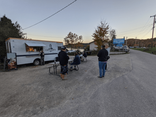 A food truck with people waiting in line on a gravel road, surrounded by trees and a setting sun.