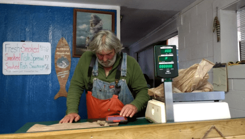 A man in overalls weighs items at a counter in a shop, with a sign displaying smoked fish products in the background.