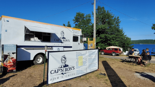 A food truck named "Cap'n Ron's Fish 'n' Chips" parked by a lake, with customers dining at a nearby table.